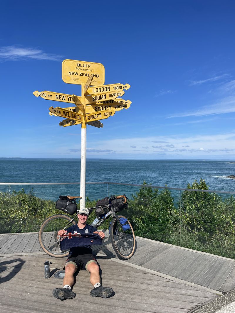 Tom Mitchell at the Bluff signpost at the southern tip of New Zealand, his loaded touring bike behind him at the end of the ride