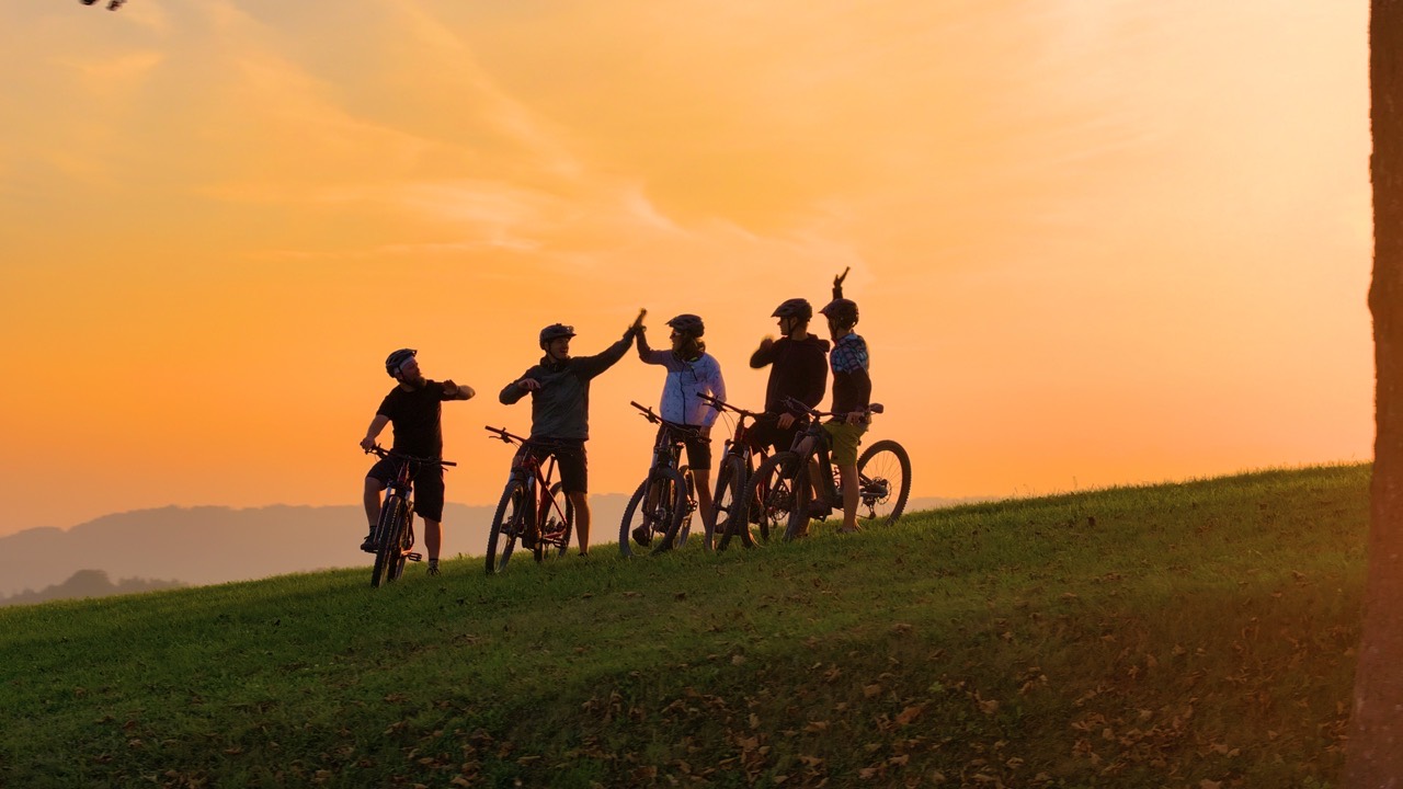 A team of five stop their bikes on a hilltop at golden hour, two of them sharing a high-five at the end of a cycling retreat day
